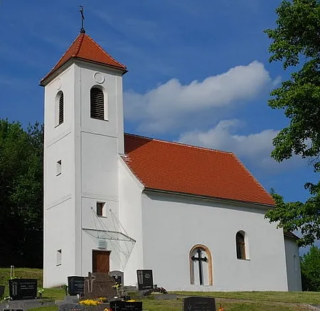 Weiße Kirche mit rotem Dach und Glockenturm, auf einem Hügel mit Friedhof gelegen.