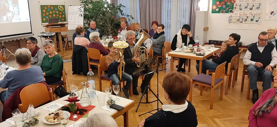 Menschen versammeln sich in einem Kirchensaal, teils musizierend, für eine Gemeindeveranstaltung.