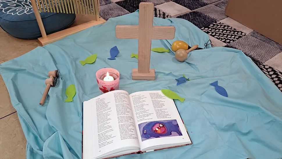 Ein Holzkreuz, offene Bibel und Kerze auf blauem Tuch, mit Filzfischen.