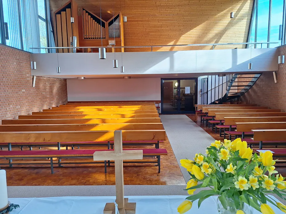 Kircheninneres mit Holzkreuz, Kirchenbänken und Orgelempore, von Sonnenlicht durchflutet.
