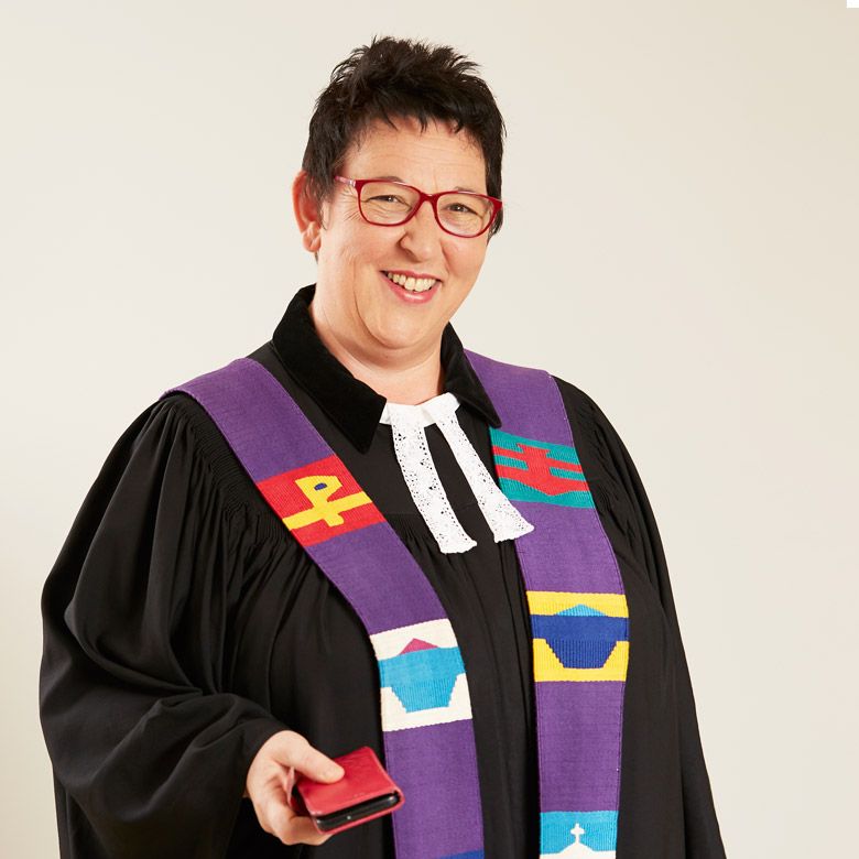 Lächelnde Frau in liturgischer Kleidung, ein kleines rotes Buch haltend.