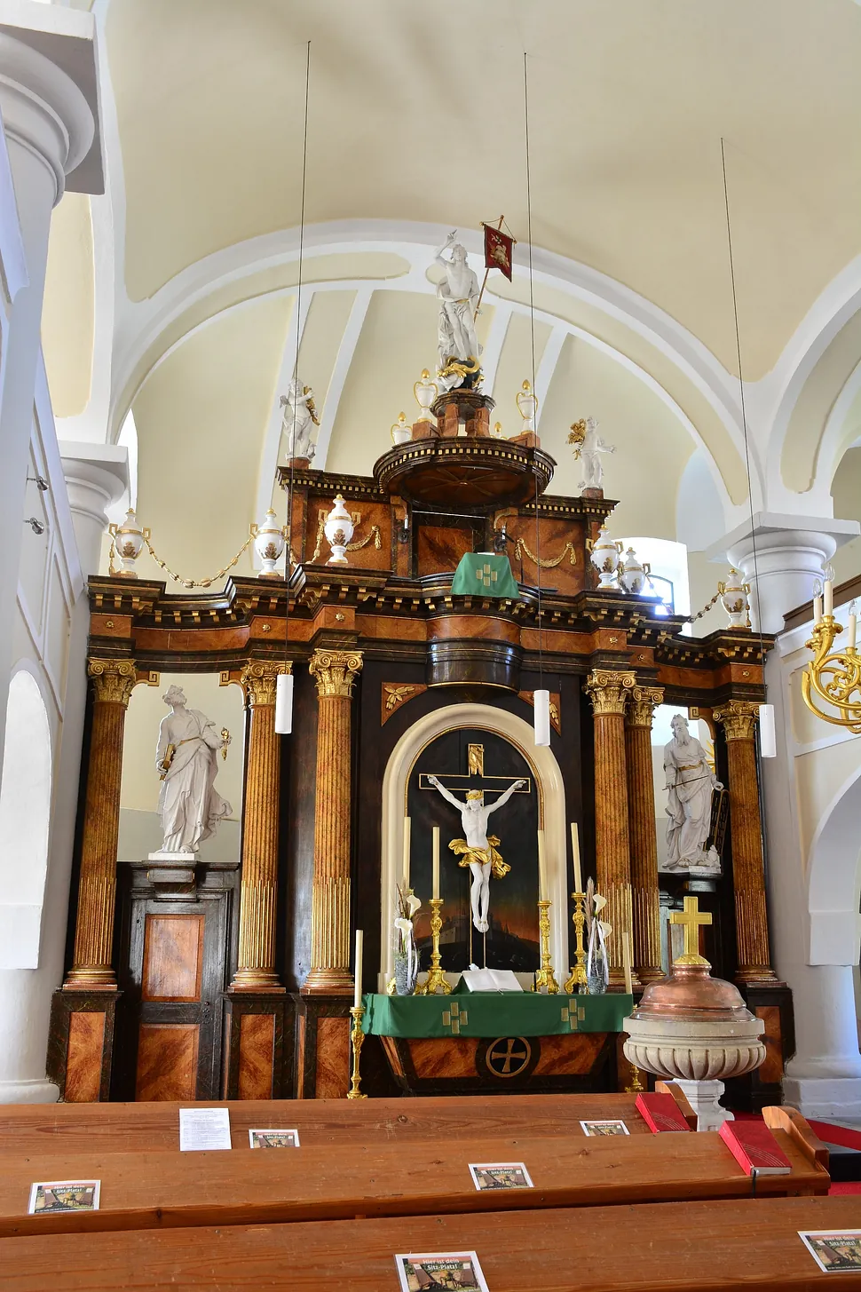 Verzierter Kirchenaltar mit Kruzifix und verschiedenen Statuen, von den Kirchenbänken aus gesehen.