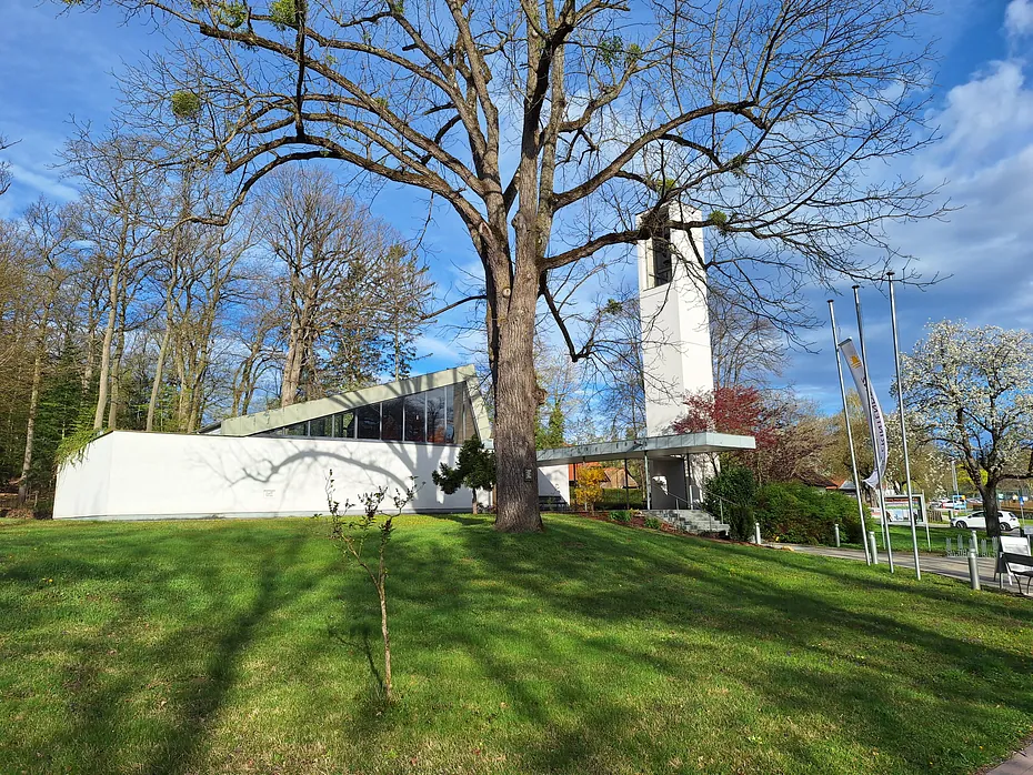 Moderne weiße Kirche mit hohem Glockenturm, umgeben von Bäumen und grünem Gras.