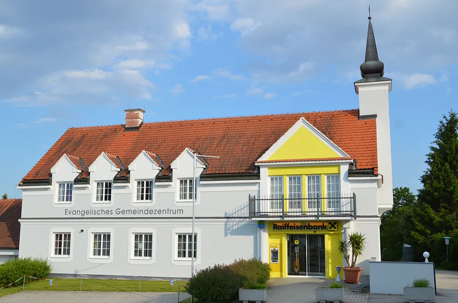 Evangelisches Gemeindezentrum mit gelber Fassade und angrenzender Bank, unter leicht bewölktem Himmel.