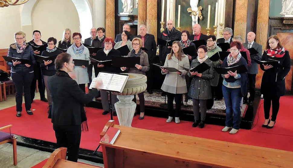 Ein Kirchenchor, mit Dirigent, singt von Notenblättern auf rotem Teppich.