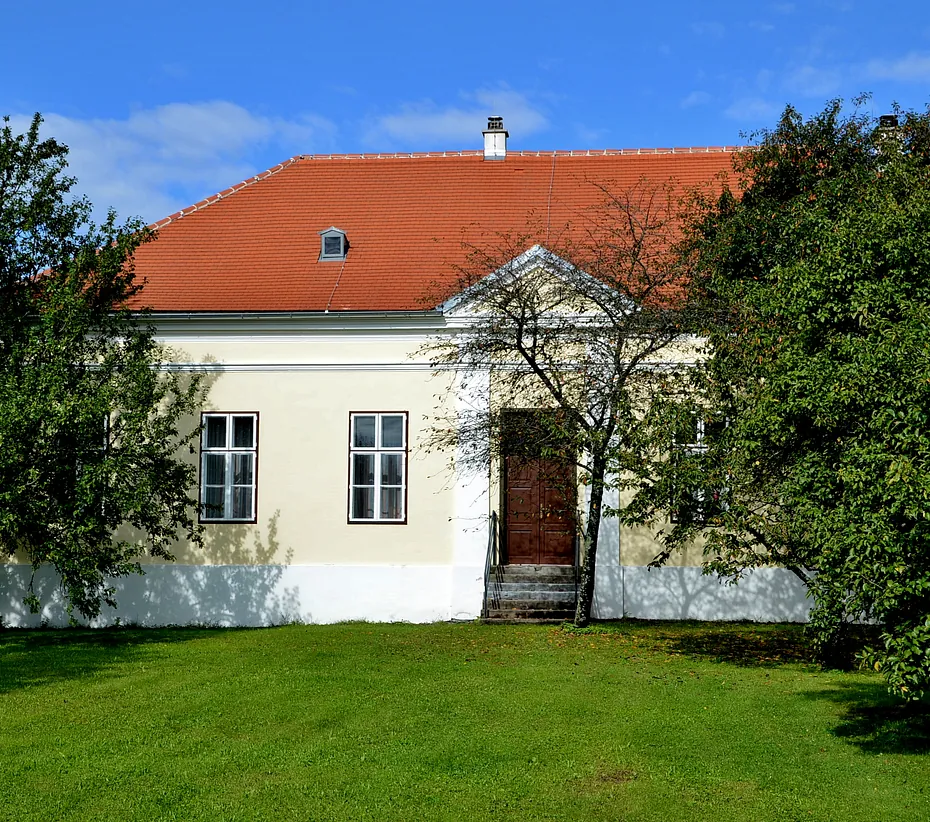 Historisches cremefarbenes Gebäude mit rotdach, umgeben von Bäumen und grünem Rasen.